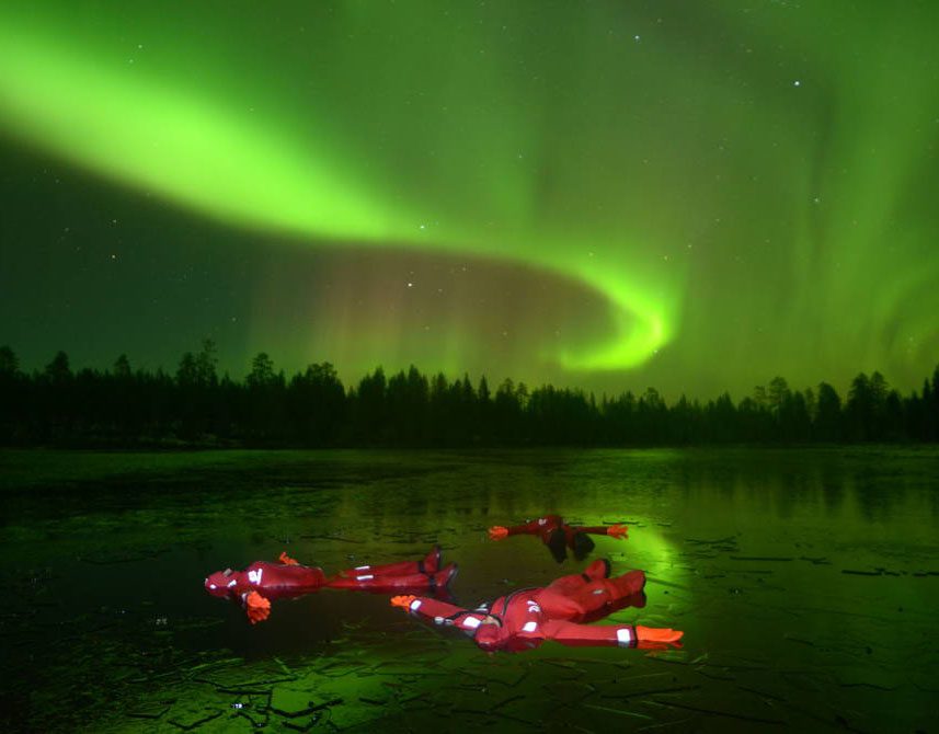 People float on a lake beneath vibrant green northern lights.