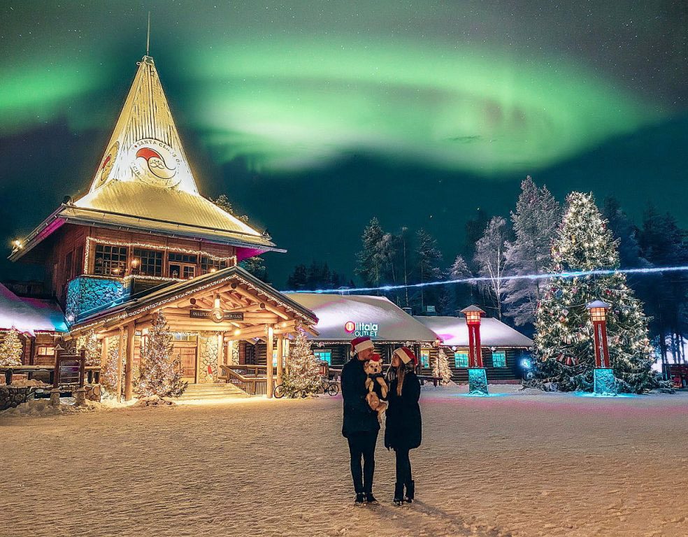 Two people stand in snowy village under northern lights.
