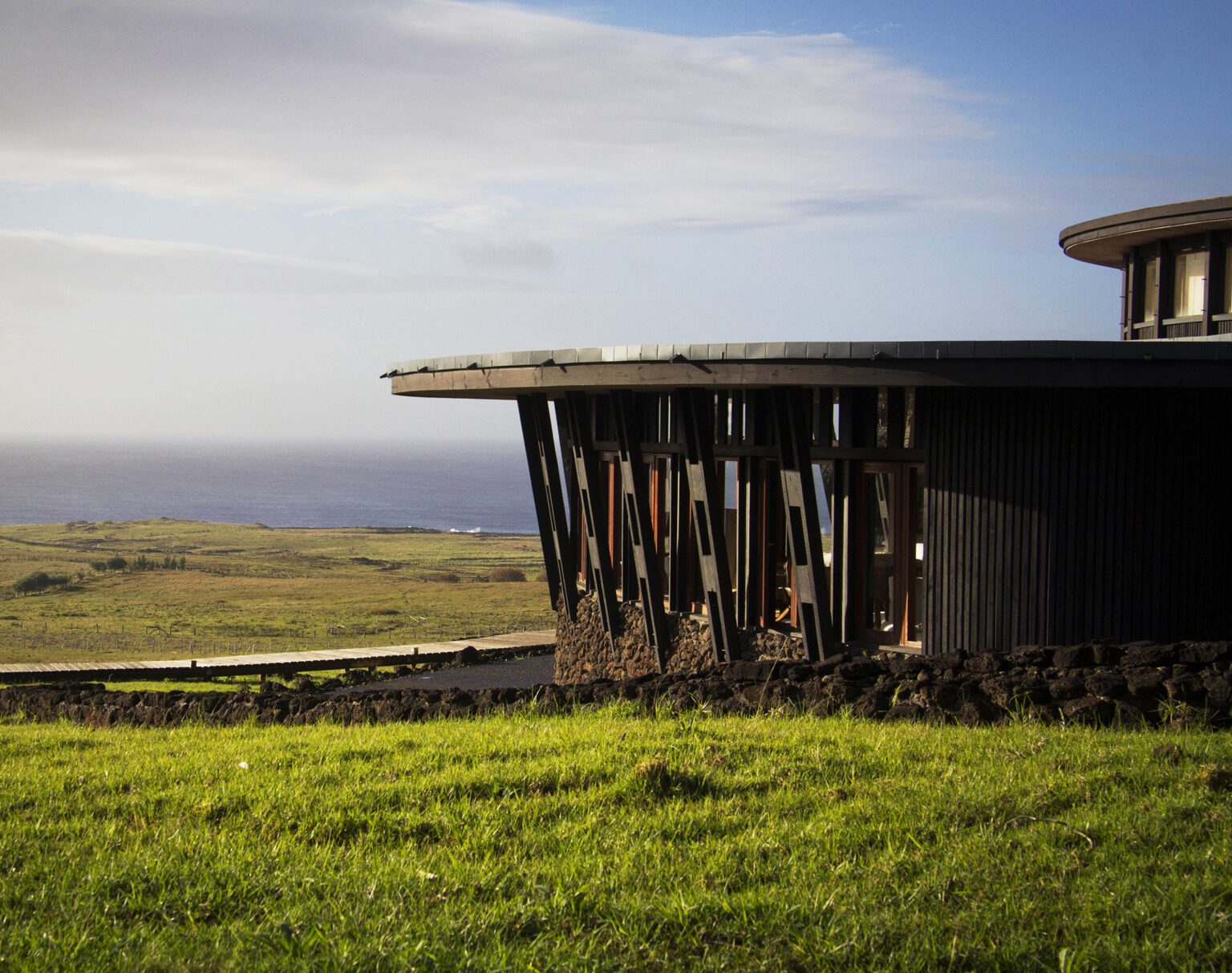 Modern cabin with wooden pillars in a grassy landscape near the sea.