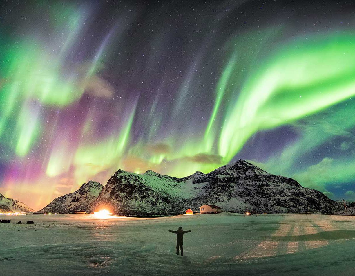 Person stands in awe beneath vibrant northern lights over snowy mountains.