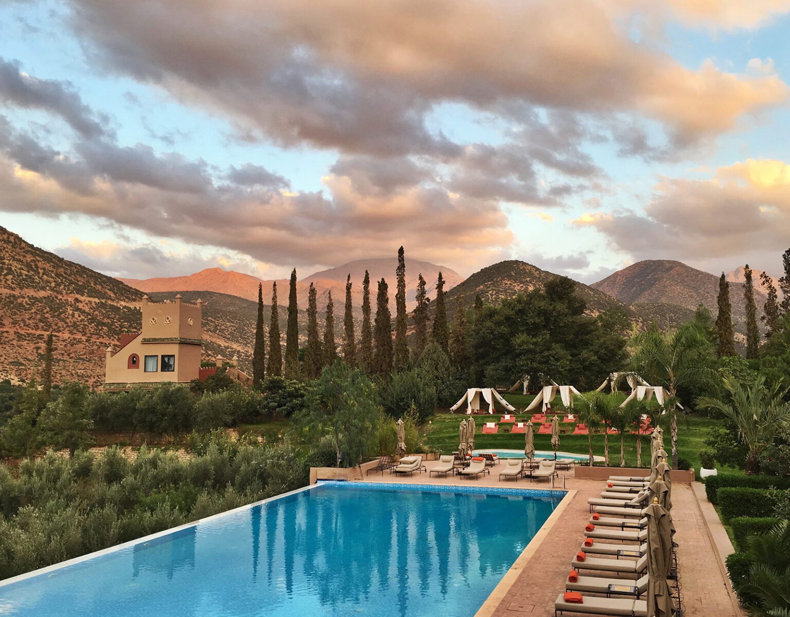 Serene poolside view with mountains and cypress trees at sunset.