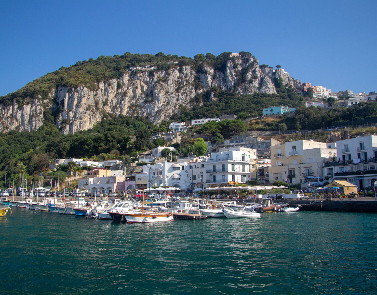 Harbor with boats and white buildings beneath a rocky cliff under a clear blue sky.