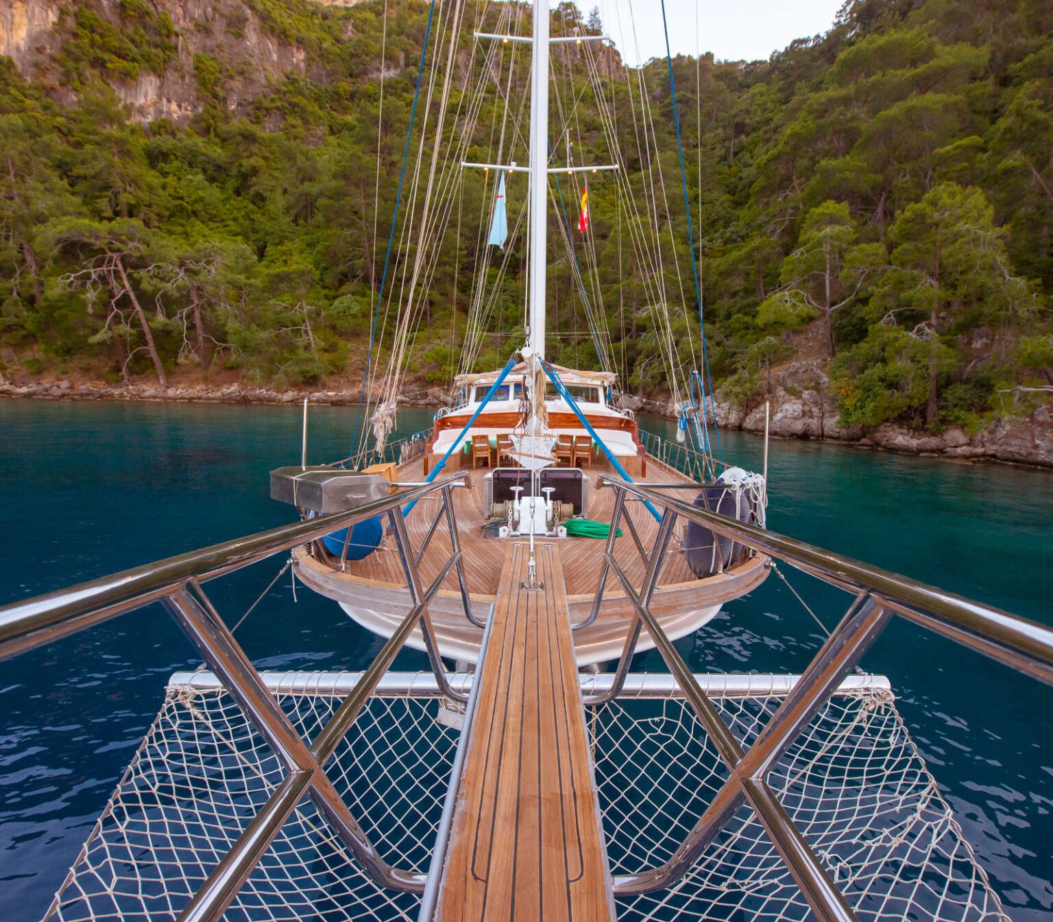View from the bow of a sailboat cruising through clear blue waters surrounded by lush greenery.