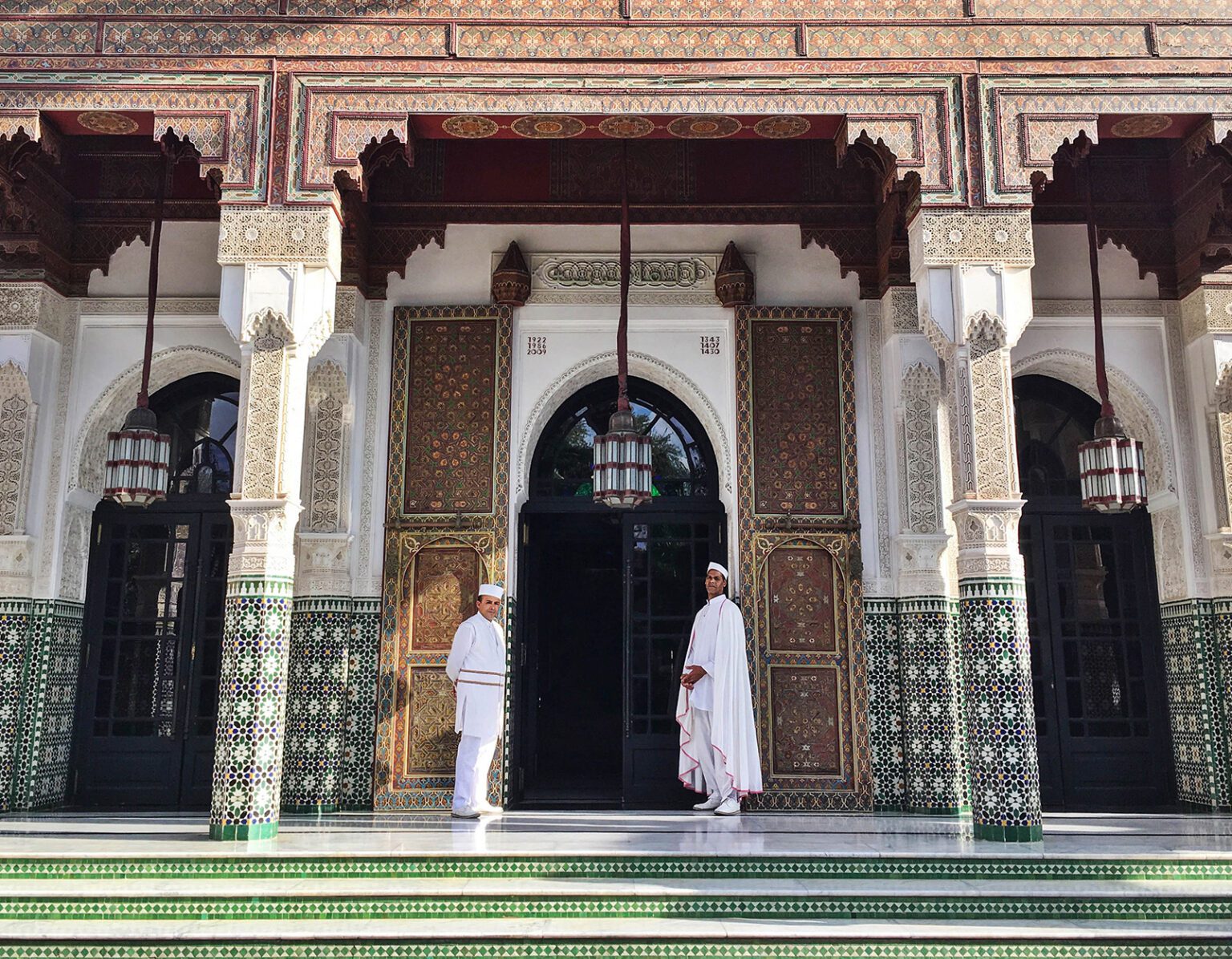 Two men in traditional attire stand outside an ornate building with intricate tile work.