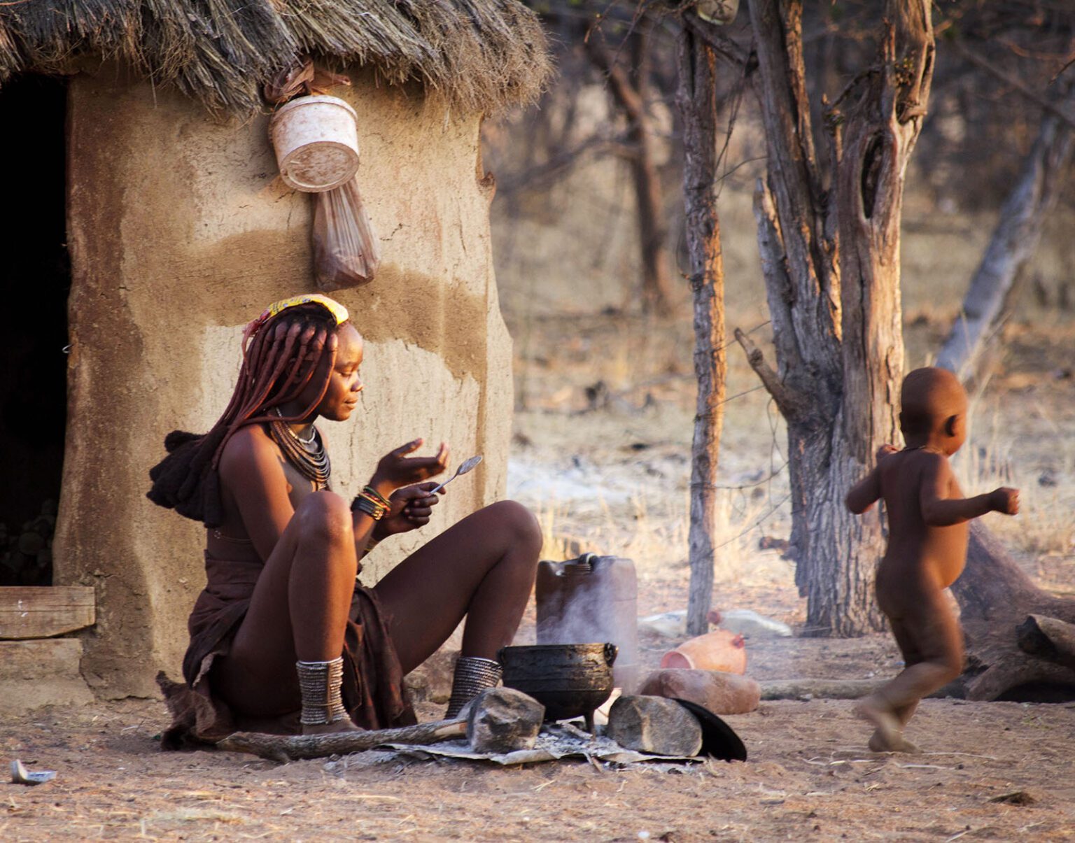 Two indigenous people sitting outside a traditional hut in a rural setting.