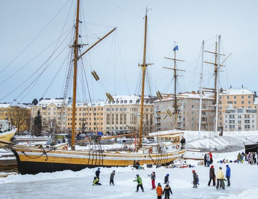 People ice skating near tall ships docked in a harbor during winter.