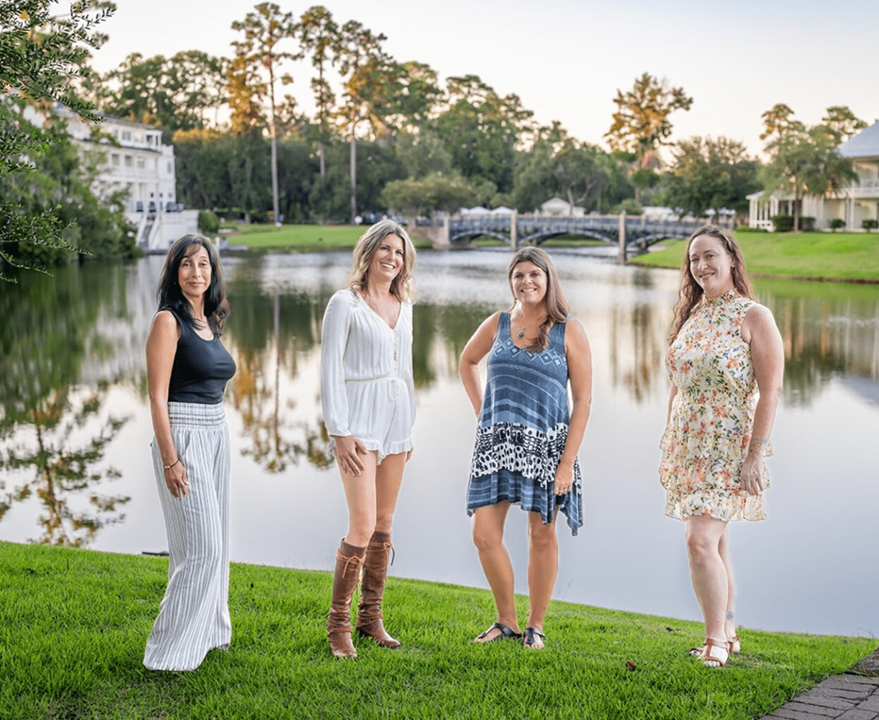 Four women standing by a lake during sunset, smiling.