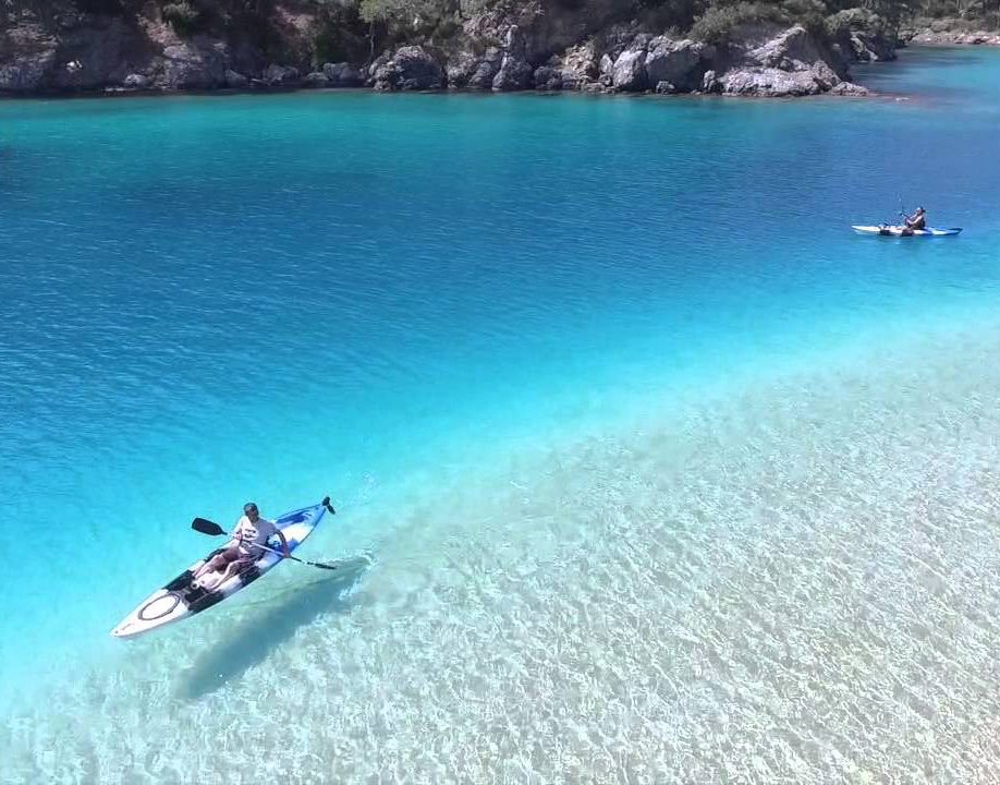 Two people kayaking on clear turquoise water near a rocky shore.
