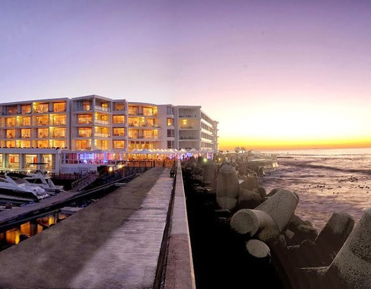 Sunset view of a waterfront promenade with illuminated buildings and rocky shore.