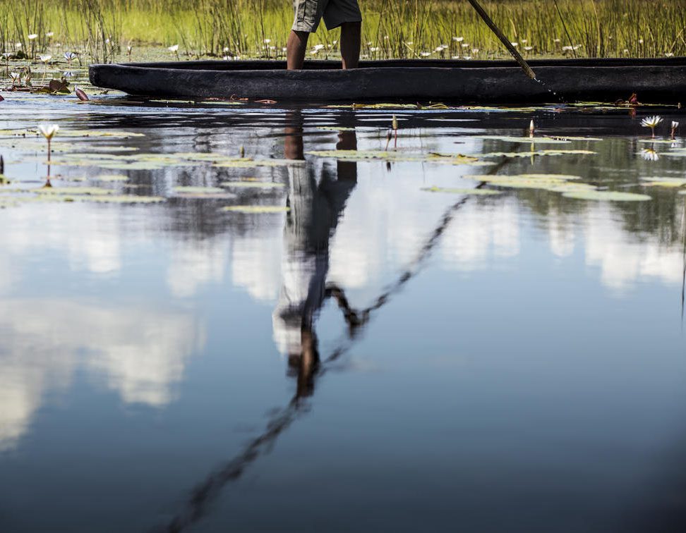 Person standing on a boat reflected in calm water with greenery.