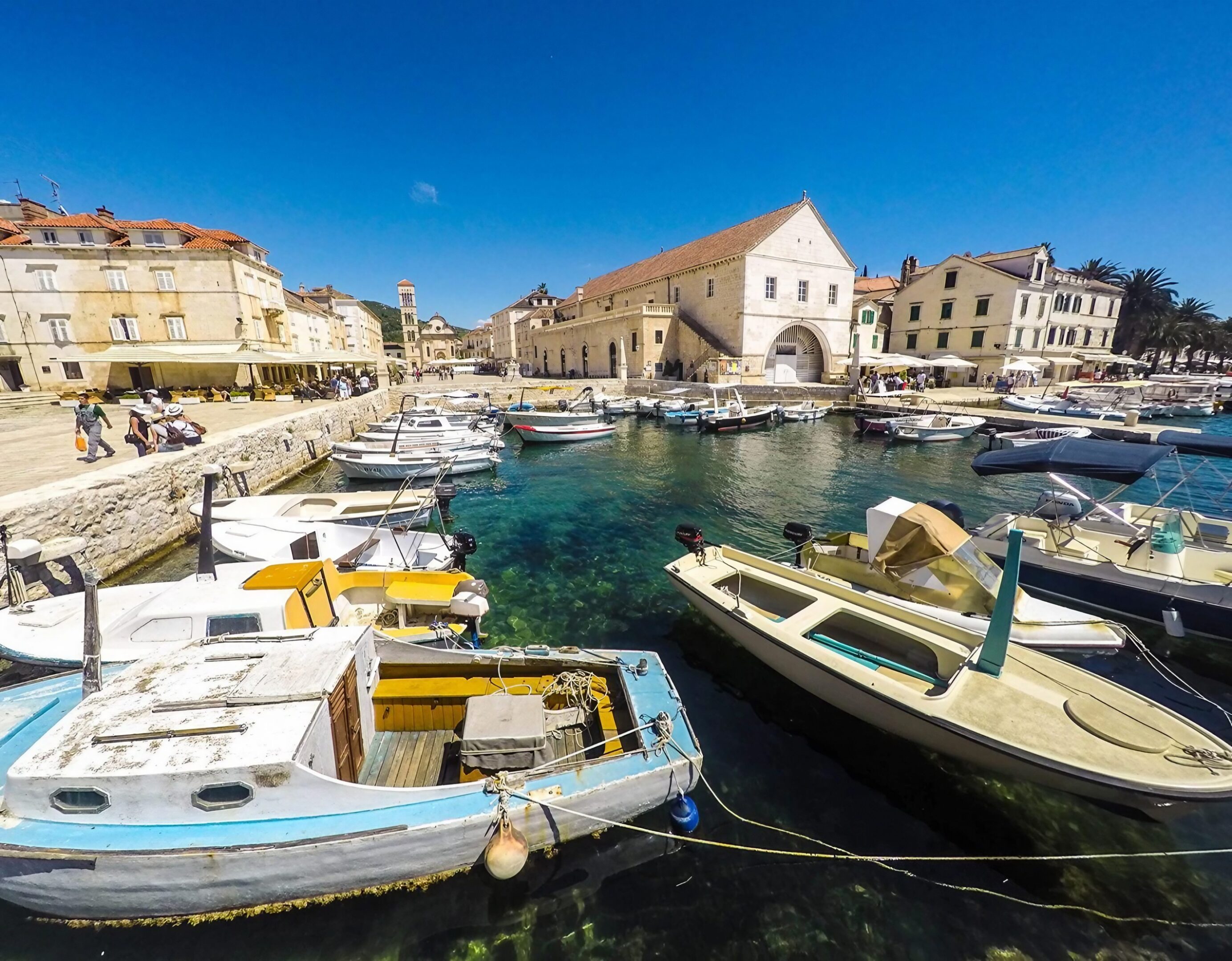 Small boats docked in a sunny Mediterranean harbor with historic buildings.