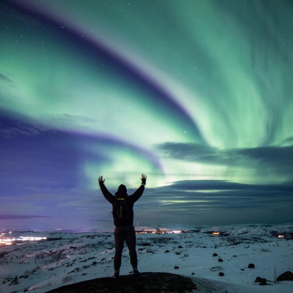 Person silhouetted against vivid green northern lights in snowy landscape.