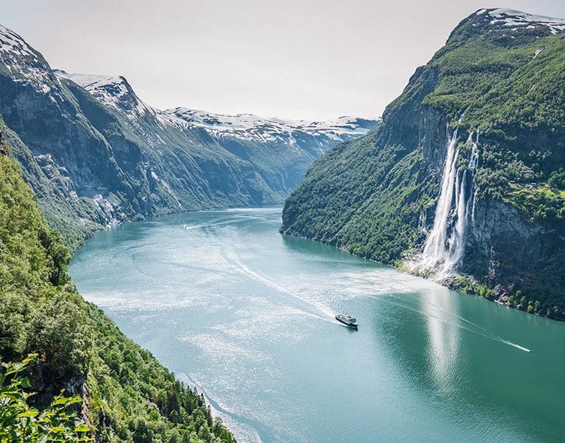 Boat cruising emerald fjord beside tall waterfall